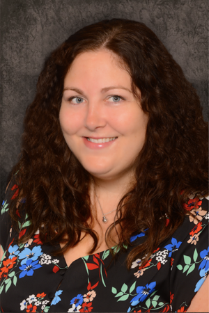 Sarah Steliga a woman, Sarah Steliga, with long curly brown hair, and wearing a black blouse with red, blue, white, and green floral patterns