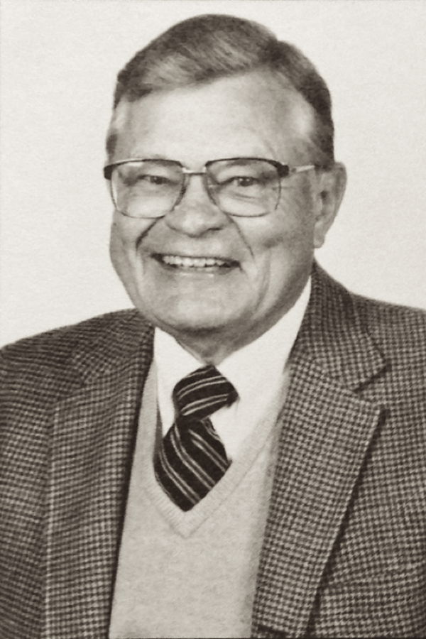 A black and white photo of a smiling man, Robert Danielson, with combed hair, greying temples, and square eyeglasses. He wears a tweed sport coat, a light colored sweater vest, a diagonally striped necktie, and a white button-down dress shirt.
