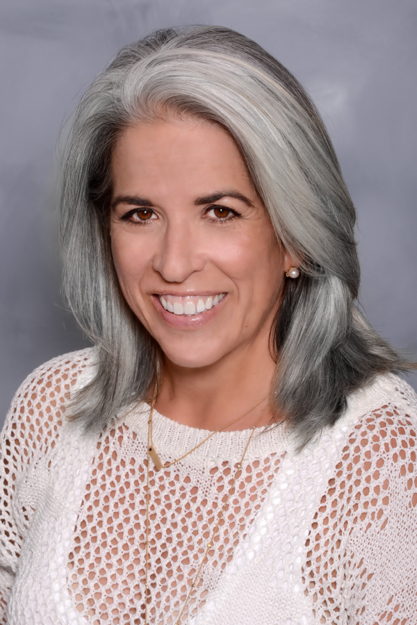 Cindi Harris, Department Co-Chair A smiling woman, Cindi Harris, with shoulder-length grey hair, wearing a white lace top, a pearl earring, and a small chain necklace.