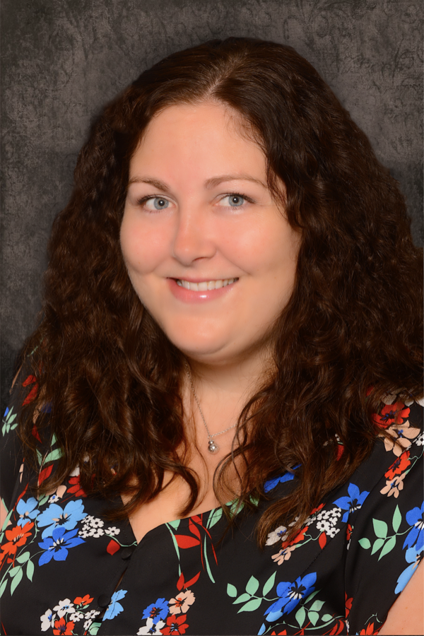 Sarah Steliga, Department Co-Chair A photo of a smiling woman, Sarah Steliga, with long, curly brown hair draping her shoulders, and wearing a black dress with a colorful floral print, and a silver beaded choker.