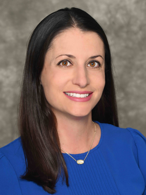 a smiling woman, Michelle Crooks, with long dark hair wearing a blue top and a small necklace, smiling against a neutral gray background