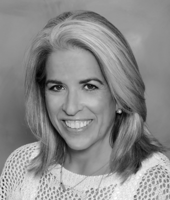 a smiling woman, Cindi Harris, with shoulder-length grey hair, wearing a white lace top, a pearl earring, and a small chain necklace