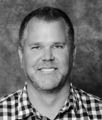 a black and white photo of a smiling man, Tate Hurvitz, with greying temples and light, greying facial hair, wearing a gingham pattern, open-collared dress shirt