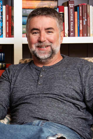 Rich Farrell Photograph of a man, Rich Farrell, with gray hair and beard sitting on a couch in front of a bookshelf filled with books.