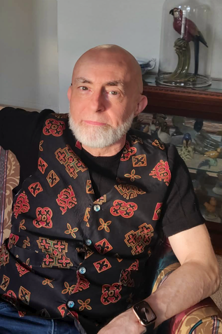 Karl Sherlock Photograph of a seated older man, Karl Sherlock, with bald head and white beard, a black short-sleeve shirt, and a colorful vest with jewel-tone designs.